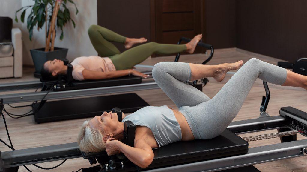 Dona estirada fent exercici de cames en una màquina reformer de Pilates, amb una altra persona practicant al fons.